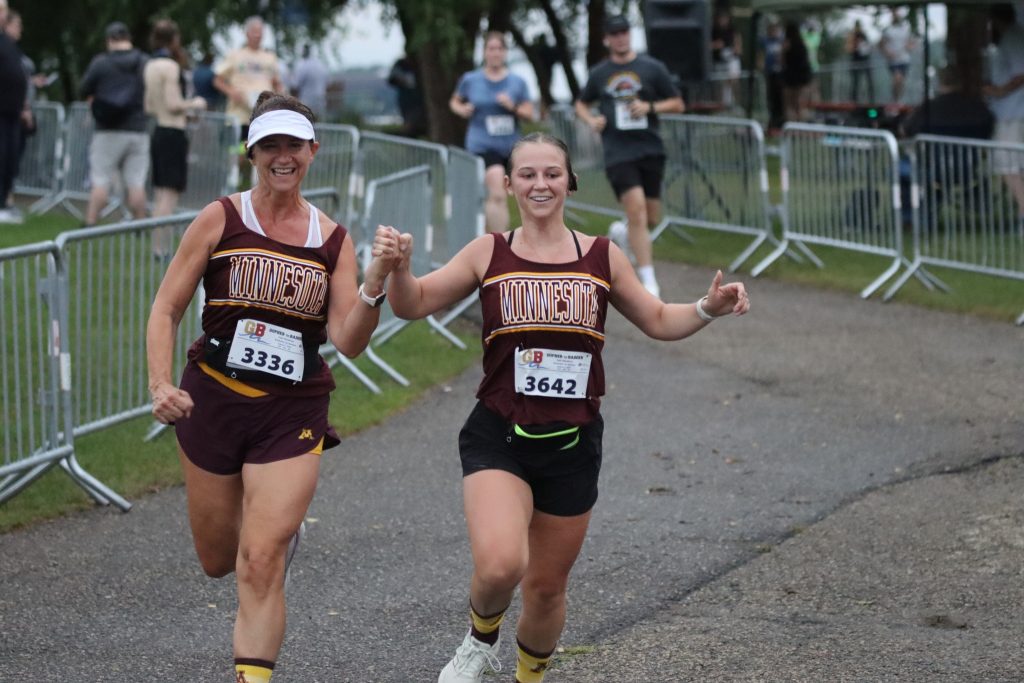 Two runners in Minnesota shirts hold hands as they run in the Gopher to Badger Half Marathon